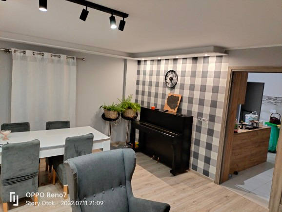 Salon z szarą tapetą w kratę, czarnym pianinem, białym stołem i szarymi krzesłami; widoczne oświetlenie punktowe na suficie.