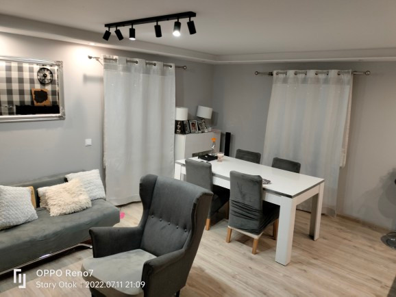 Jasne wnętrze pokoju dziennego z szarą sofą, fotelem typu uszak, białym stołem jadalnym i zasłonami w tle, oświetlone czarnym reflektorem sufitowym.