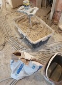 Mieszanie zaprawy w czarnym pojemniku przy użyciu mieszadła elektrycznego, widoczne kable elektryczne, otwarty worek z zaprawą i wiadro na podłodze w trakcie prac budowlanych.