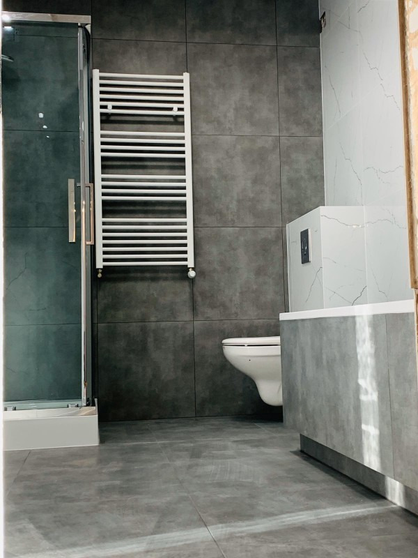 Nowoczesna łazienka z szarymi płytkami, białym grzejnikiem drabinkowym na ścianie, kabiną prysznicową ze szklanymi drzwiami i podwieszaną toaletą.