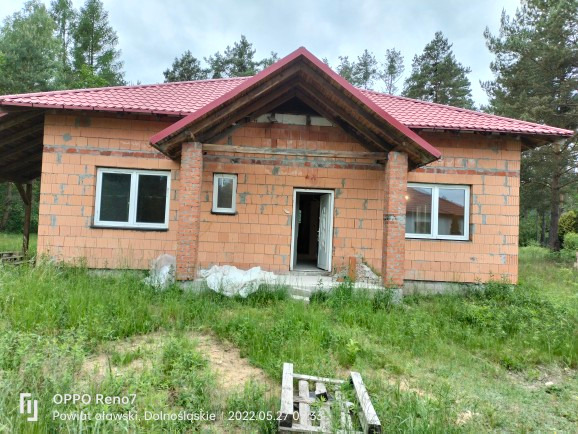 Dom jednorodzinny w stanie surowym otwartym, z czerwoną dachówką i białymi oknami, otoczony zielenią, widok z przodu na niezakończony budynek.