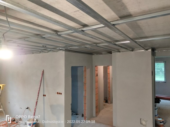 Wnętrze w trakcie prac wykończeniowych: metalowa konstrukcja sufitu podwieszanego, ściany po wstępnym szpachlowaniu, widoczne otwory drzwiowe z surową cegłą, poziomica oparta o ścianę, instalacja...