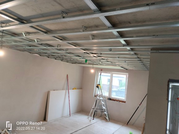 Metalowa konstrukcja sufitowa w trakcie montażu, widoczne profile i zawiesia, w tle ściany przygotowane do dalszych prac wykończeniowych, drabina oparta o parapet okna.