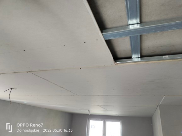 Widok na sufit w trakcie montażu, widoczne metalowe profile konstrukcyjne i częściowo zamontowane płyty, z widocznymi śladami po wkrętach.