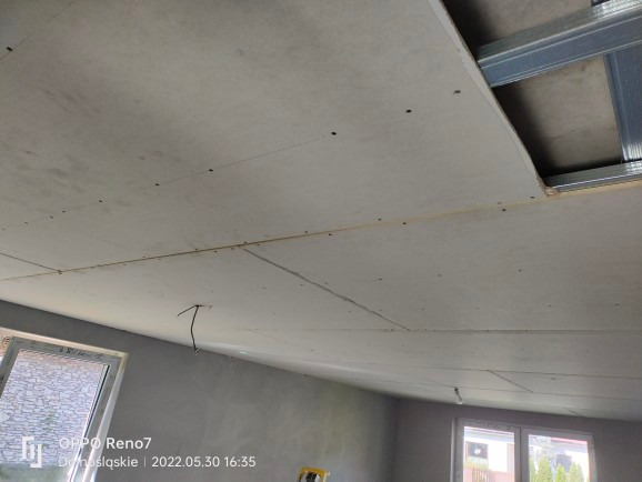 Widok na sufit z zamontowanymi płytami gipsowo-kartonowymi, widoczne metalowe profile konstrukcyjne i wystające przewody elektryczne.