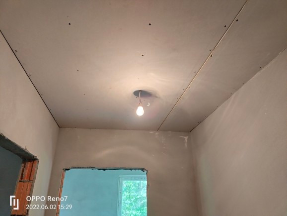 Wnętrze w trakcie remontu: widok na sufit z zamontowaną płytą, widoczne łączenia płyt i otwór na oświetlenie, ściany przygotowane do dalszych prac wykończeniowych, surowy otwór drzwiowy z cegłami.