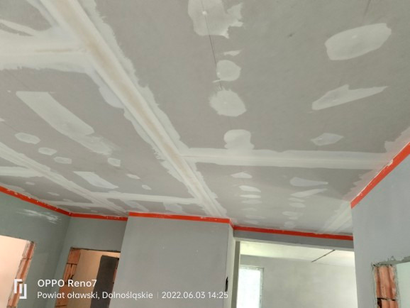 Niedokończony sufit z płyt gipsowo-kartonowych, widoczne łączenia i miejsca po wkrętach wymagające wykończenia, zabezpieczone pomarańczową taśmą malarską na obwodzie, w tle surowe ściany i otwory...