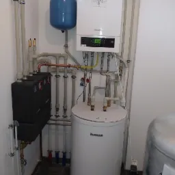 Kompletna instalacja grzewcza z piecem gazowym Buderus, zasobnikiem CWU i naczyniem przeponowym w kotłowni z szarą podłogą i białymi ścianami, widoczne rury miedziane i plastikowe.