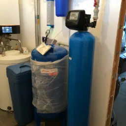 Niebieskie filtry do wody w piwnicy, jeden owinięty folią, widoczne rury i zawory, obok piec grzewczy i odpływ w podłodze.