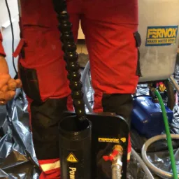 Instalator w czerwonym kombinezonie używa urządzenia Powerflushing Filter marki Fernox do czyszczenia instalacji, widoczny zbiornik i przewody na folii ochronnej.