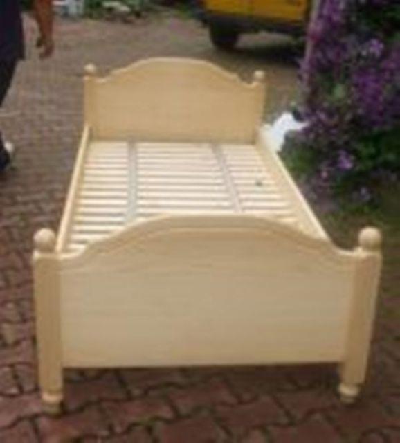 Beżowe łóżko dziecięce z drewnianym stelażem, sfotografowane na zewnątrz na tle kostki brukowej, z widocznym fragmentem żółtego samochodu i fioletowych kwiatów w tle.