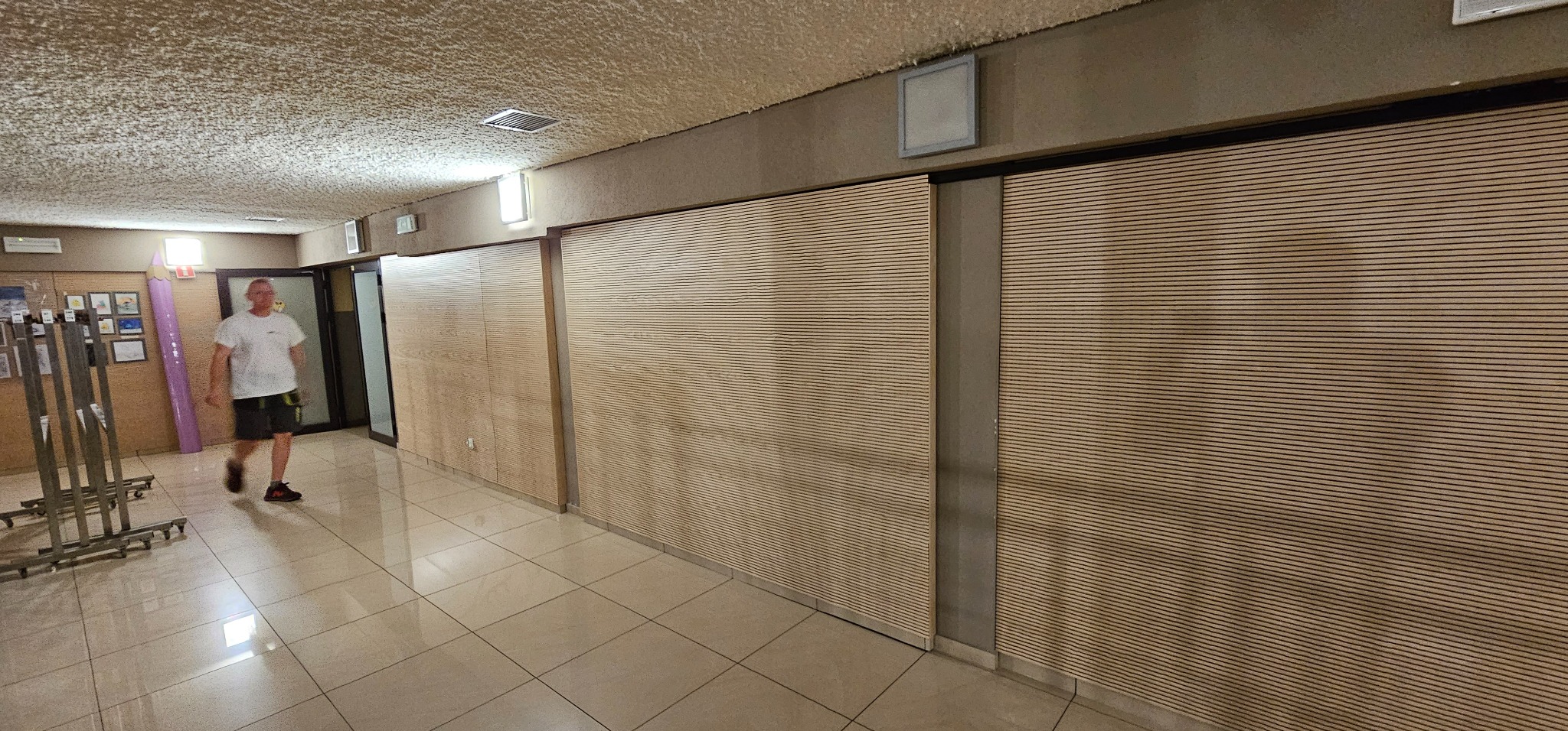 Korytarz z wykończonymi ścianami z paneli drewnianych i sufitem z fakturą baranka, w tle mężczyzna przechodzący obok metalowego wieszaka na kółkach i dekoracji w kształcie kredki.