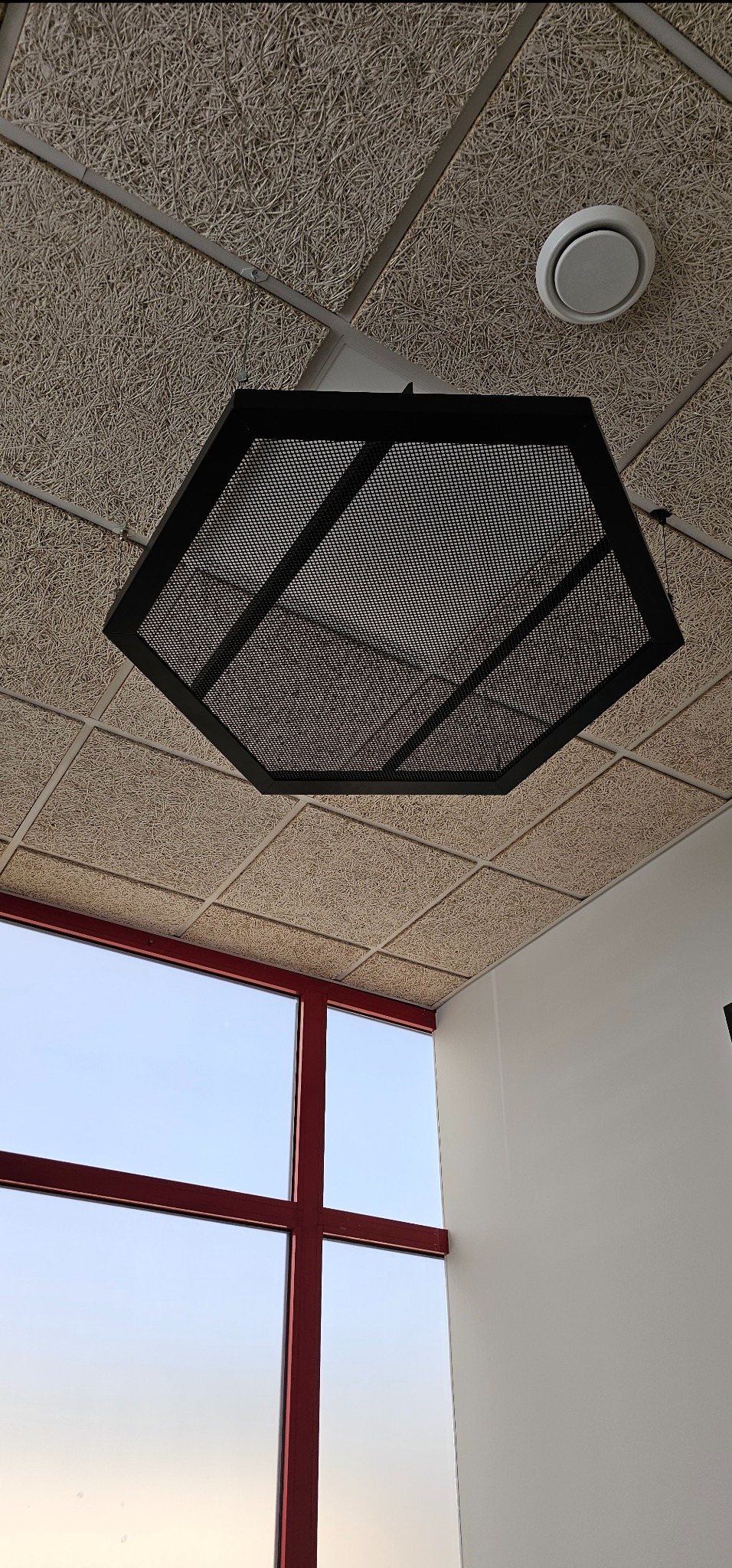 Sufit z płyt drewnopodobnych z sześciokątną lampą rastrową, widok na okno z czerwonymi ramami.