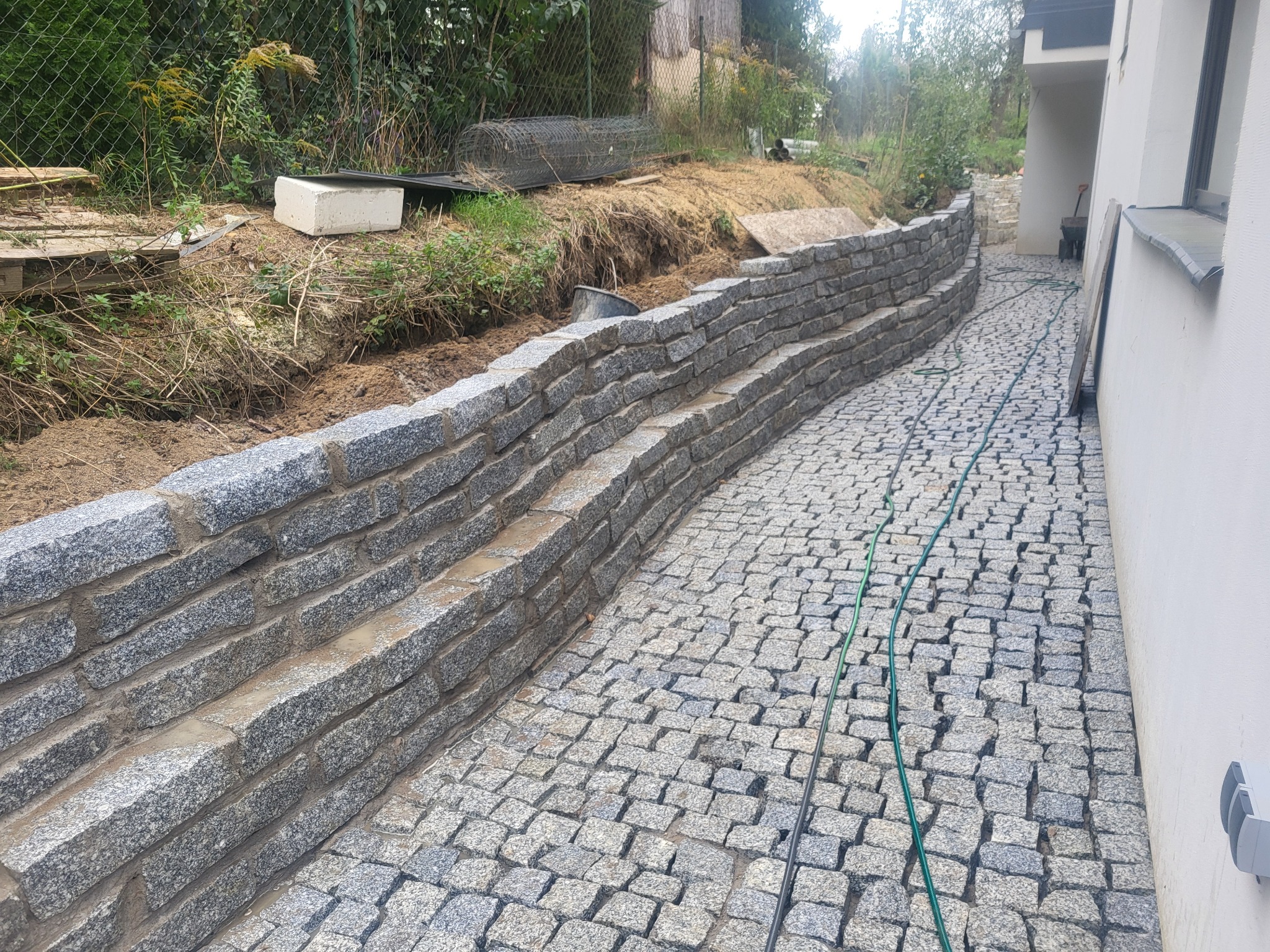 Granitowa kostka brukowa ułożona wzdłuż ściany budynku, tworząca ścieżkę z niskim murem oporowym. Widoczny fragment ogrodu z siatką ogrodzeniową w tle.