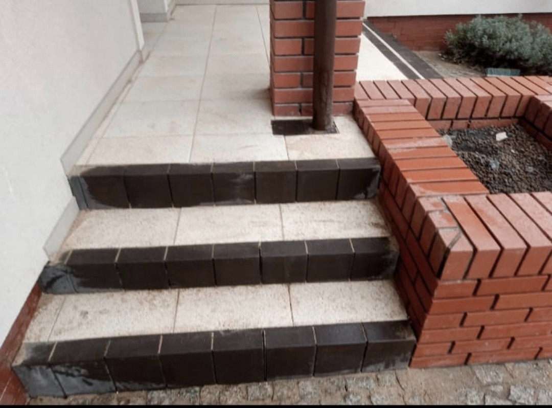 Wykończone schody wejściowe z płytek i obrzeży z kostki, obok mur z cegły. Widoczny fragment elewacji i rynna. Ujęcie z dołu, podkreślające detale wykończenia.