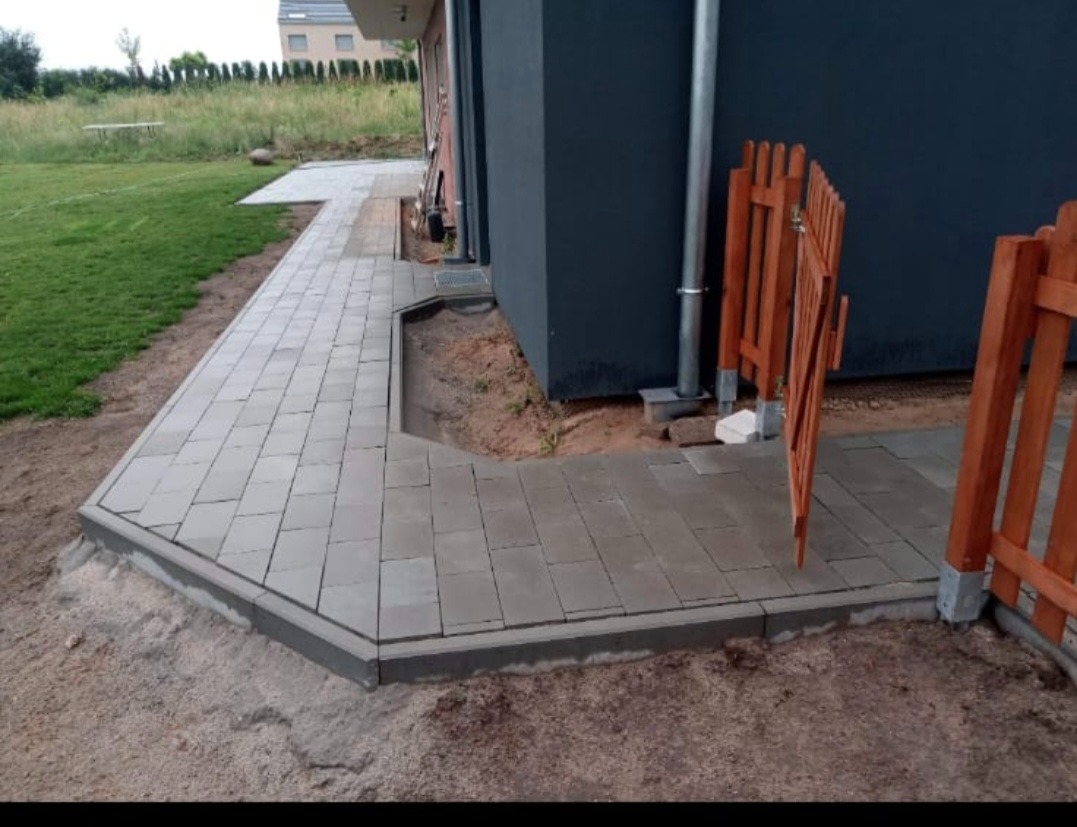 Szara kostka brukowa ułożona wzdłuż budynku z drewnianym płotem, widok z boku, świeżo po ułożeniu, z widocznym piaskiem i fragmentem trawnika.