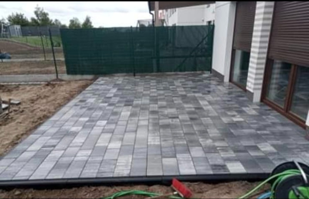 Ułożona szara kostka brukowa na tarasie przydomowym, widok z niskiej perspektywy, ogrodzenie z siatki w tle, częściowo zasłonięte okna z brązowymi ramami.