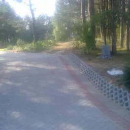 Ułożona kostka brukowa w kolorze szarym z czerwoną obwódką, prowadząca przez teren zalesiony. Widoczne betonowe ażurowe elementy obrzeża.