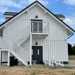 Biały dom z zewnętrznymi schodami, czarnym dachem i drzwiami, otoczony trawnikiem i drzewami pod błękitnym niebem z chmurami.