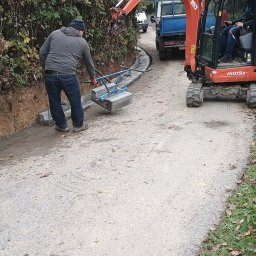 FU Polkop Wawrzynek Tomasz - Układanie krawężników wzdłuż drogi gruntowej z użyciem mini koparki i specjalistycznego podnośnika. Prace drogowe w toku, widoczny pracownik i maszyny.