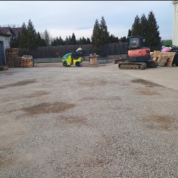 FU Polkop Wawrzynek Tomasz - Żółty walec drogowy w trakcie pracy na żwirowej powierzchni, obok koparka Kubota i palety. W tle budynek i ogrodzenie z tujami. Utwardzanie terenu w toku.