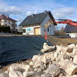 FU Polkop Wawrzynek Tomasz - Czerwona koparka Kubota na placu budowy, obok sterta jasnych kamieni i czarna geowłóknina. W tle domy w różnym stanie ukończenia i błękitne niebo.