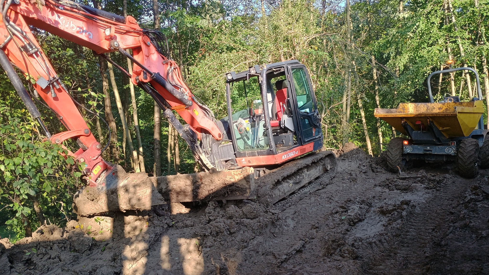 Pomarańczowa koparka Kubota KX080-4 w trakcie prac ziemnych na błotnistym terenie leśnym, z żółtą wywrotką w tle, w słoneczny dzień.