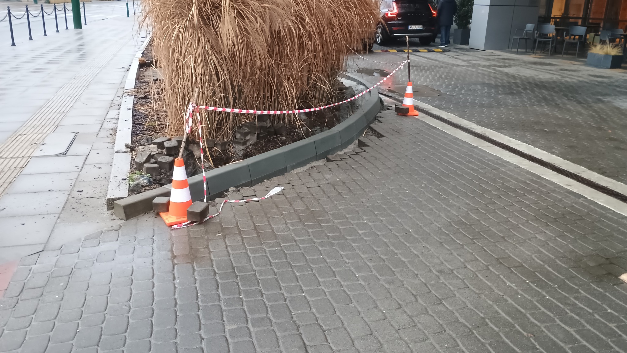Uszkodzona nawierzchnia chodnika z kostki brukowej w Mogilanach, zabezpieczona taśmą i pachołkami. Widoczne rozkopane fragmenty i wystające elementy.