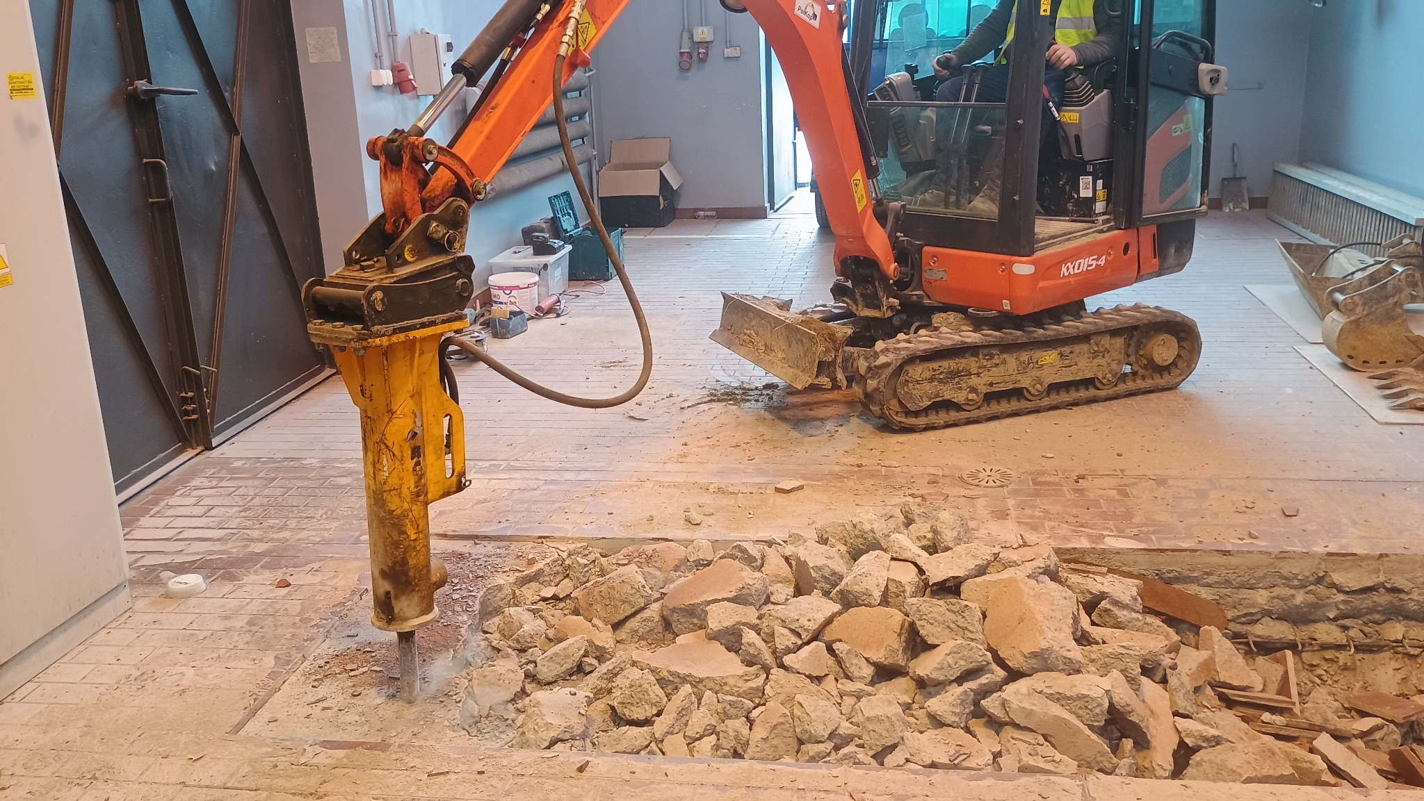 Pomarańczowa minikoparka z młotem hydraulicznym rozbija posadzkę wewnątrz budynku. Widoczne gruzowisko i fragmenty instalacji. Operator w kabinie.