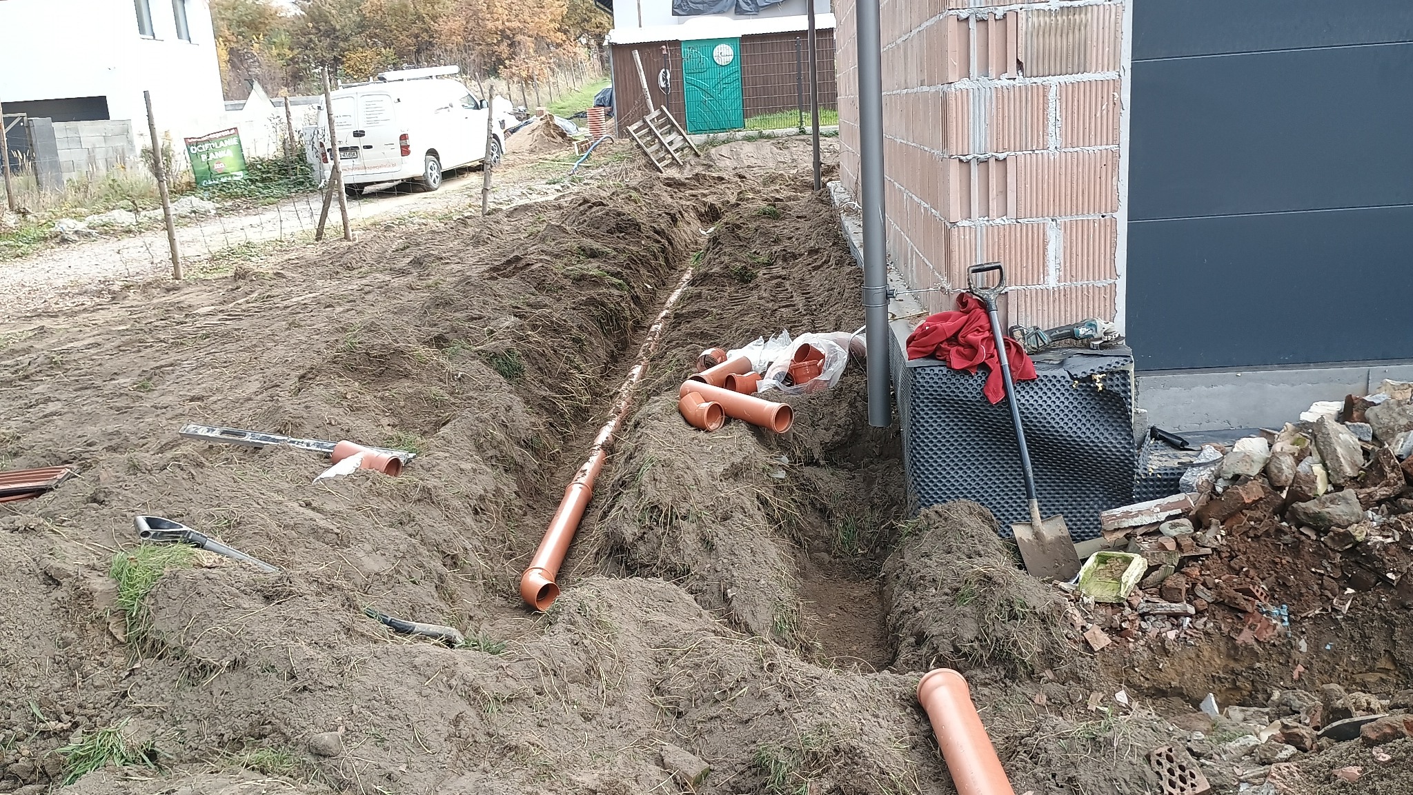 Wykopany rów z ułożonymi rurami kanalizacyjnymi z tworzywa sztucznego obok fundamentu budynku. Widoczne narzędzia i folia kubełkowa.