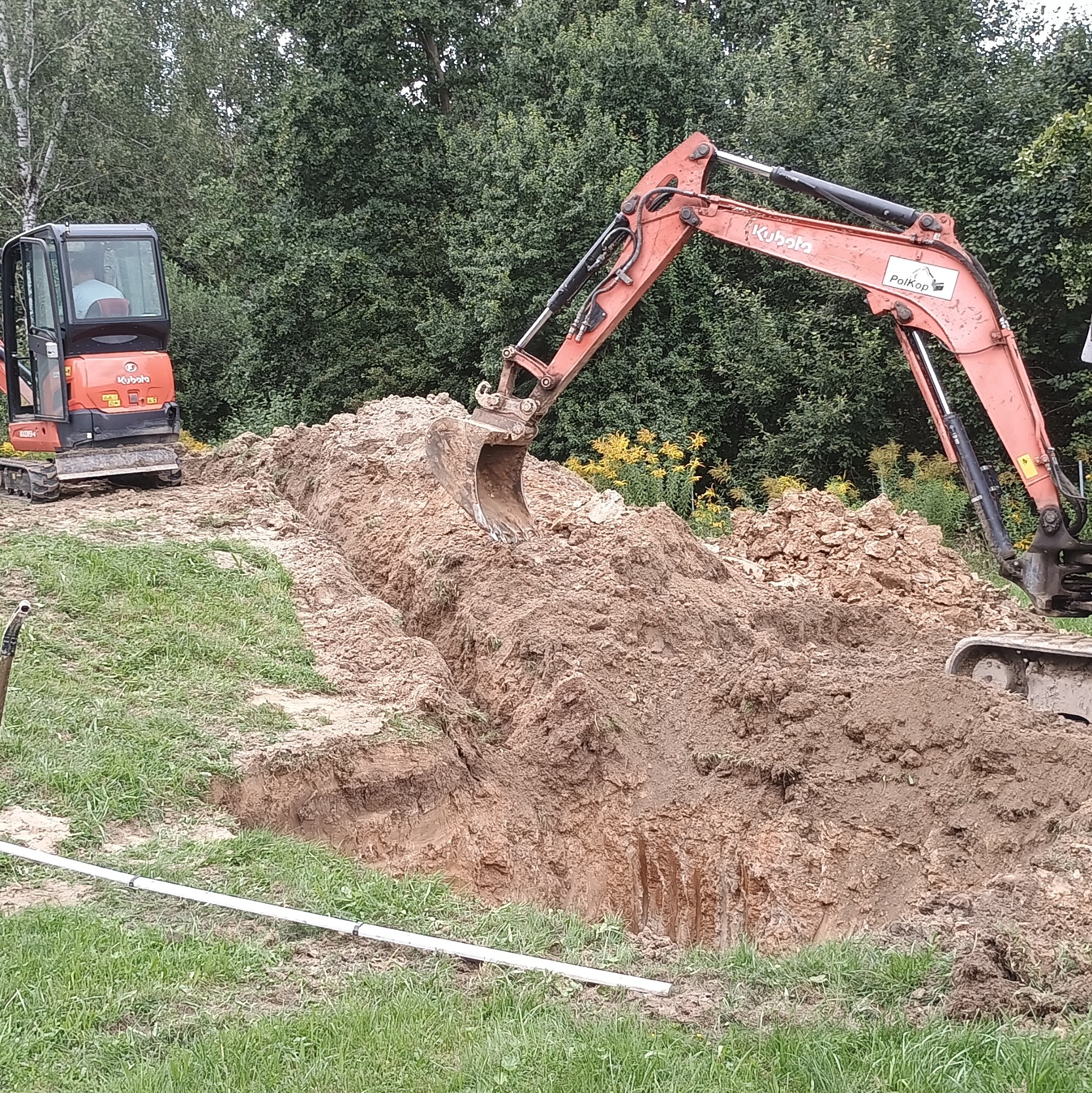 Pomarańczowa koparka Kubota w trakcie wykopu pod instalacje na zielonym terenie, widoczne rury. Praca w toku, z bliska, z drzewami w tle. Wykopana ziemia.