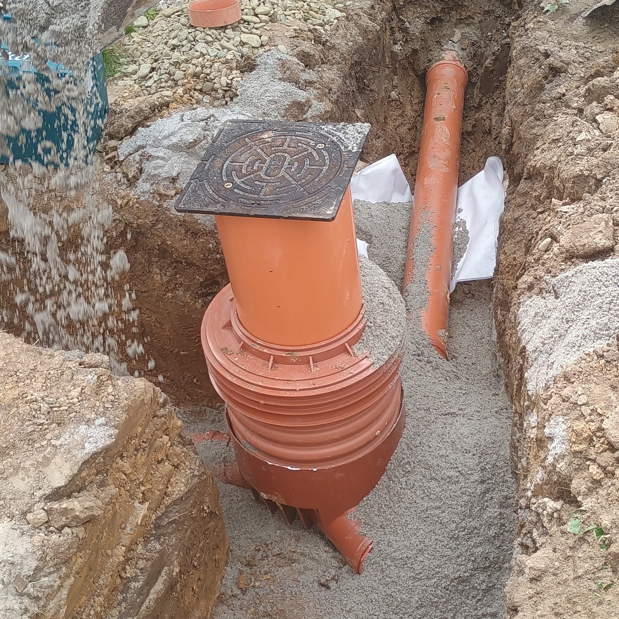 Pomarańczowa studzienka kanalizacyjna z żeliwną pokrywą w wykopie, obsypana piaskiem, z widocznymi rurami. W tle ziemia i resztki gruzu.