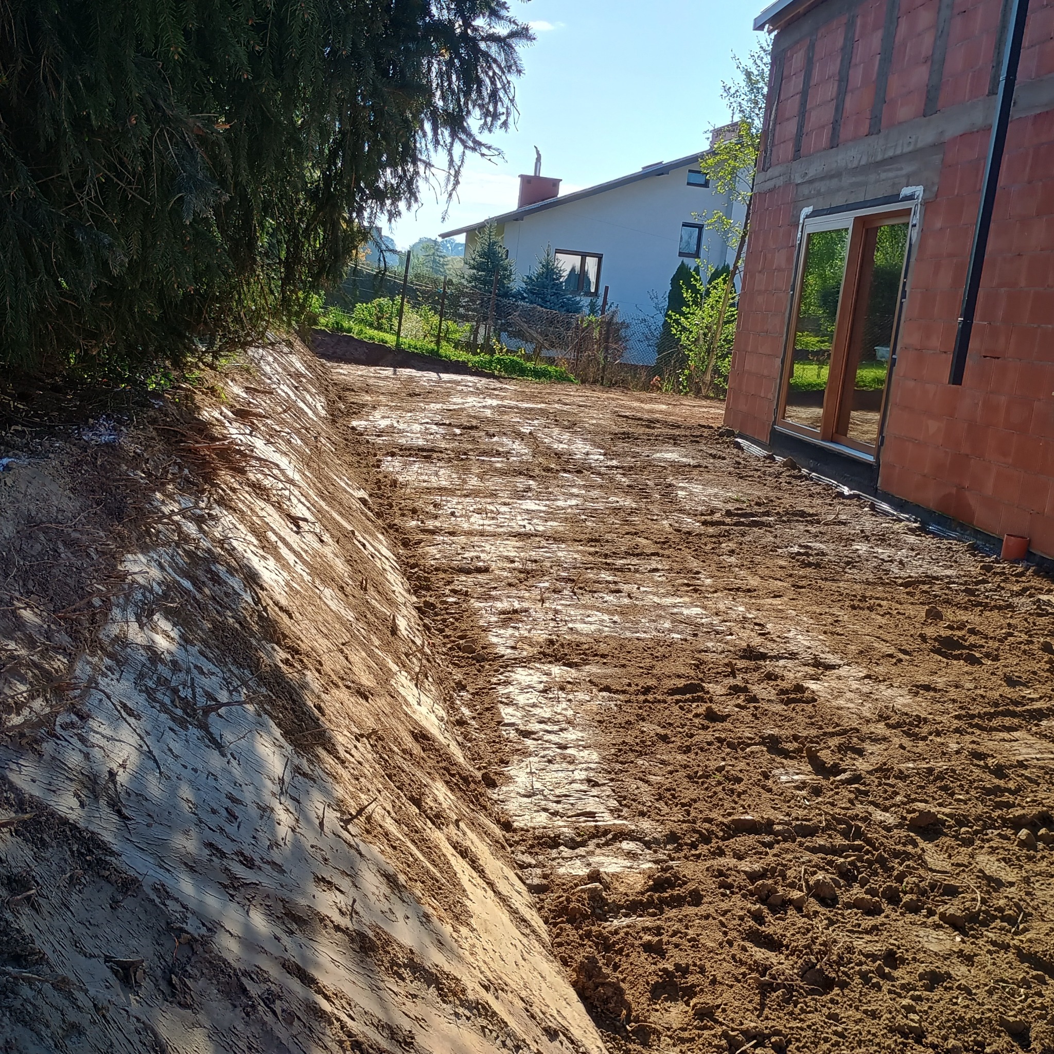 Przygotowany teren pod budowę domu jednorodzinnego; widoczne ślady po pracach ziemnych i fundamenty w surowym stanie, cegły, drzewo.