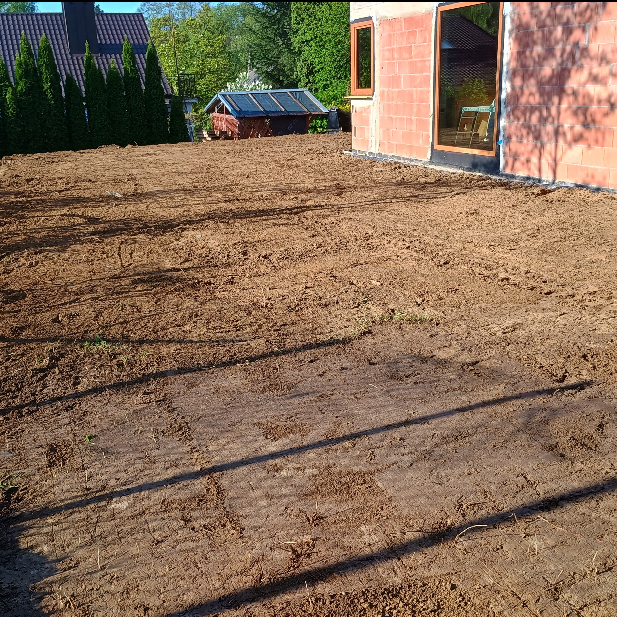 Przygotowany, wyrównany teren pod budowę domu jednorodzinnego. Widoczne ślady po maszynach budowlanych. W tle budynek w trakcie budowy i zieleń.
