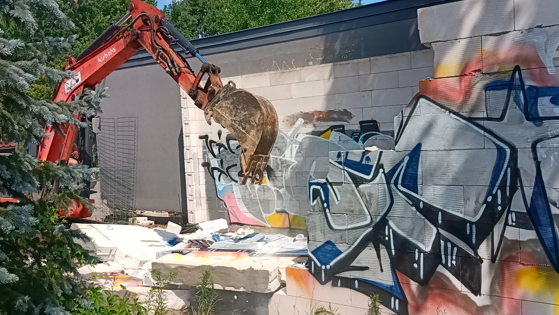 Czerwona koparka Kubota kruszy graffiti pokryty mur z betonu komórkowego. Widoczne gruz i zieleń. Praca w toku, częściowo zasłonięta przez drzewo.