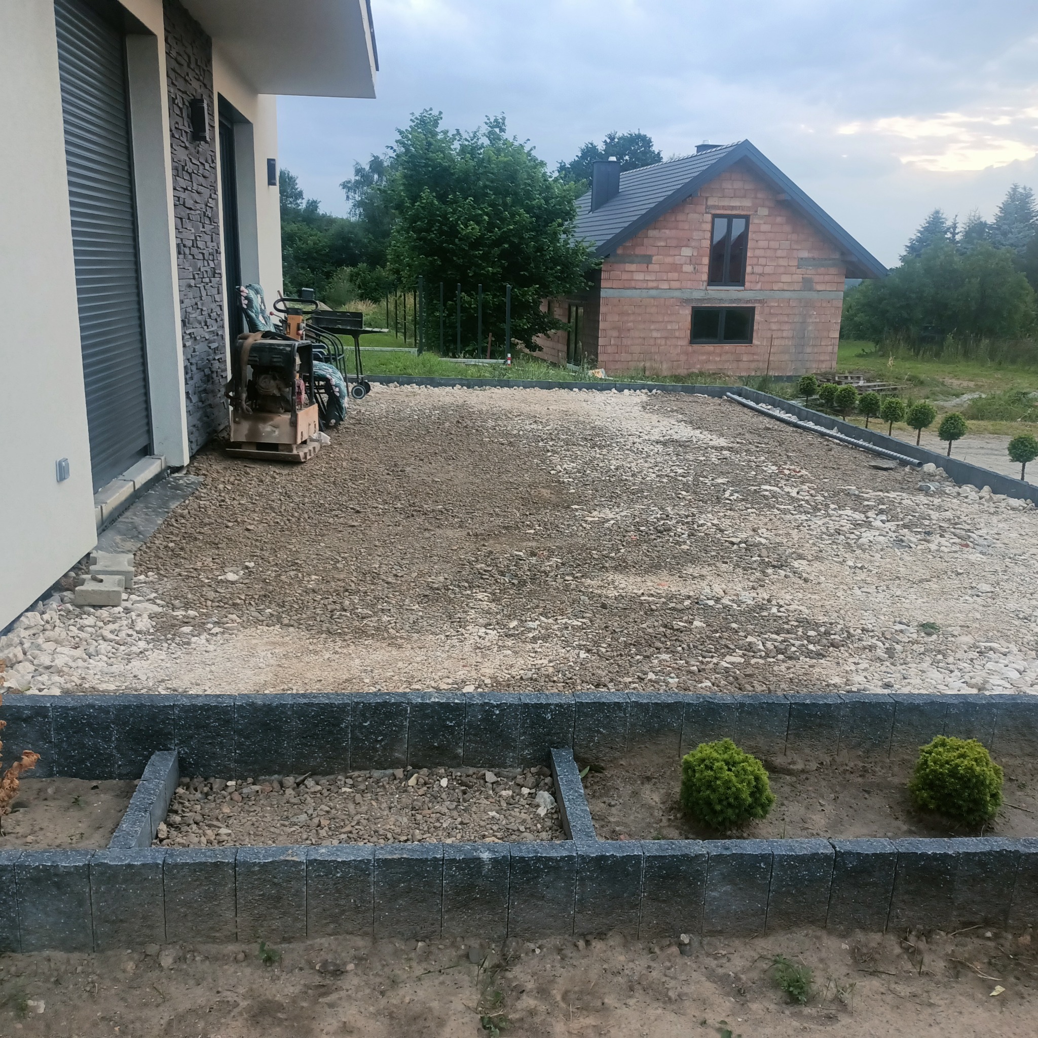 Przygotowanie podłoża pod kostkę brukową z obrzeżami. Widoczny zagęszczarka, częściowo wybrukowana powierzchnia i budynek w tle. Obrzeża z kostki betonowej.