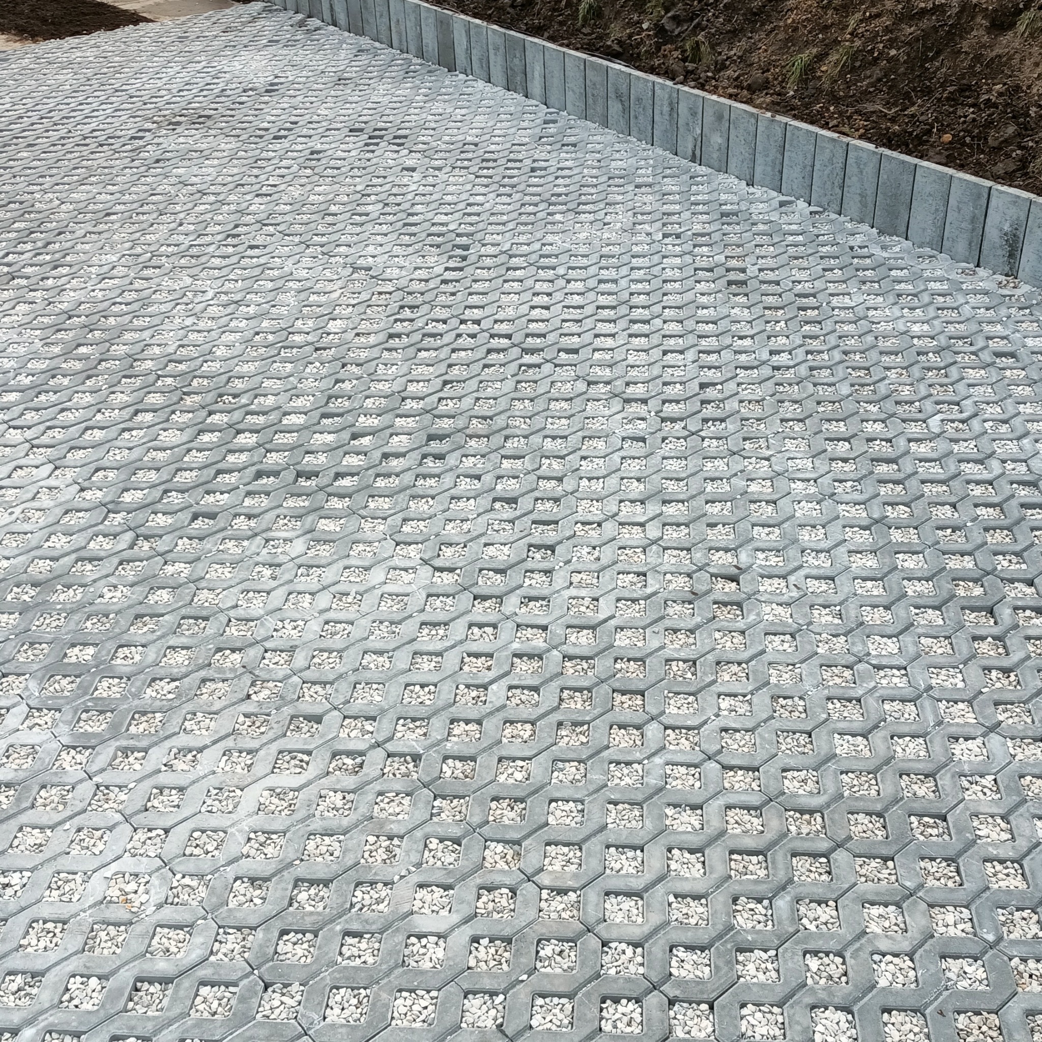 Szara ażurowa nawierzchnia z wypełnieniem z białego grysu, obrzeża z szarej kostki. Widok z góry, ukośnie. Utwardzona droga z przepuszczalną nawierzchnią.