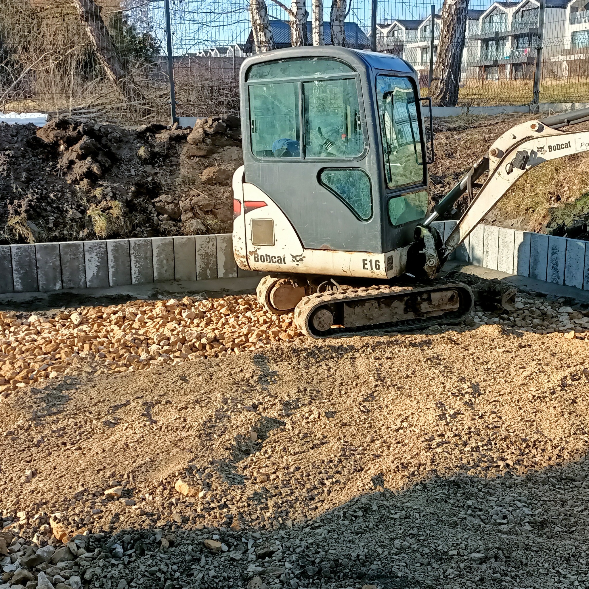 Mini koparka Bobcat E16 na placu budowy z ułożonymi obrzeżami i wysypanym kruszywem. W tle widać pryzmę ziemi i budynki mieszkalne.