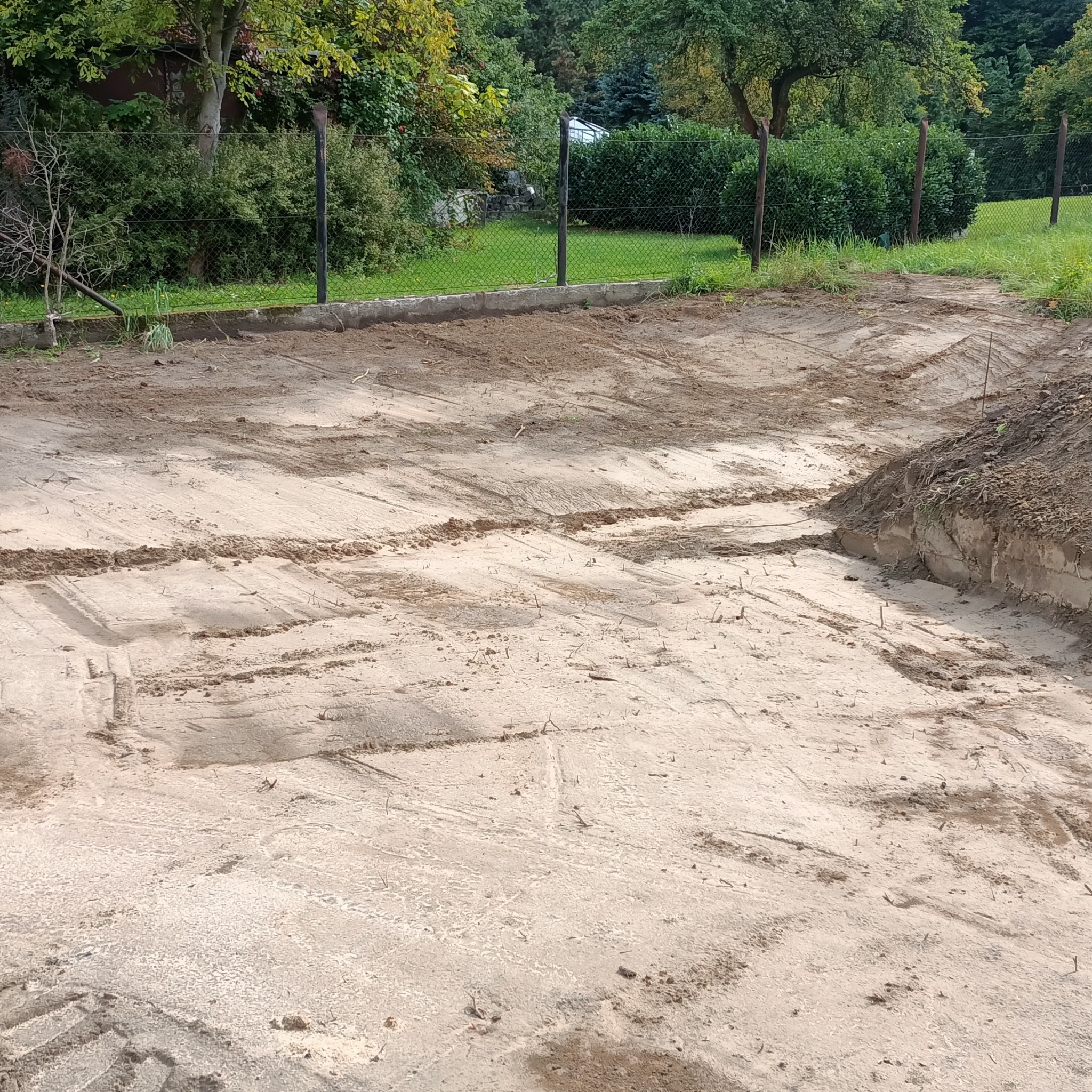 Przygotowany teren pod budowę, widoczne ślady maszyn. Grunt wyrównany, ogrodzenie w tle. Etap robót ziemnych, gotowy do dalszych prac budowlanych.