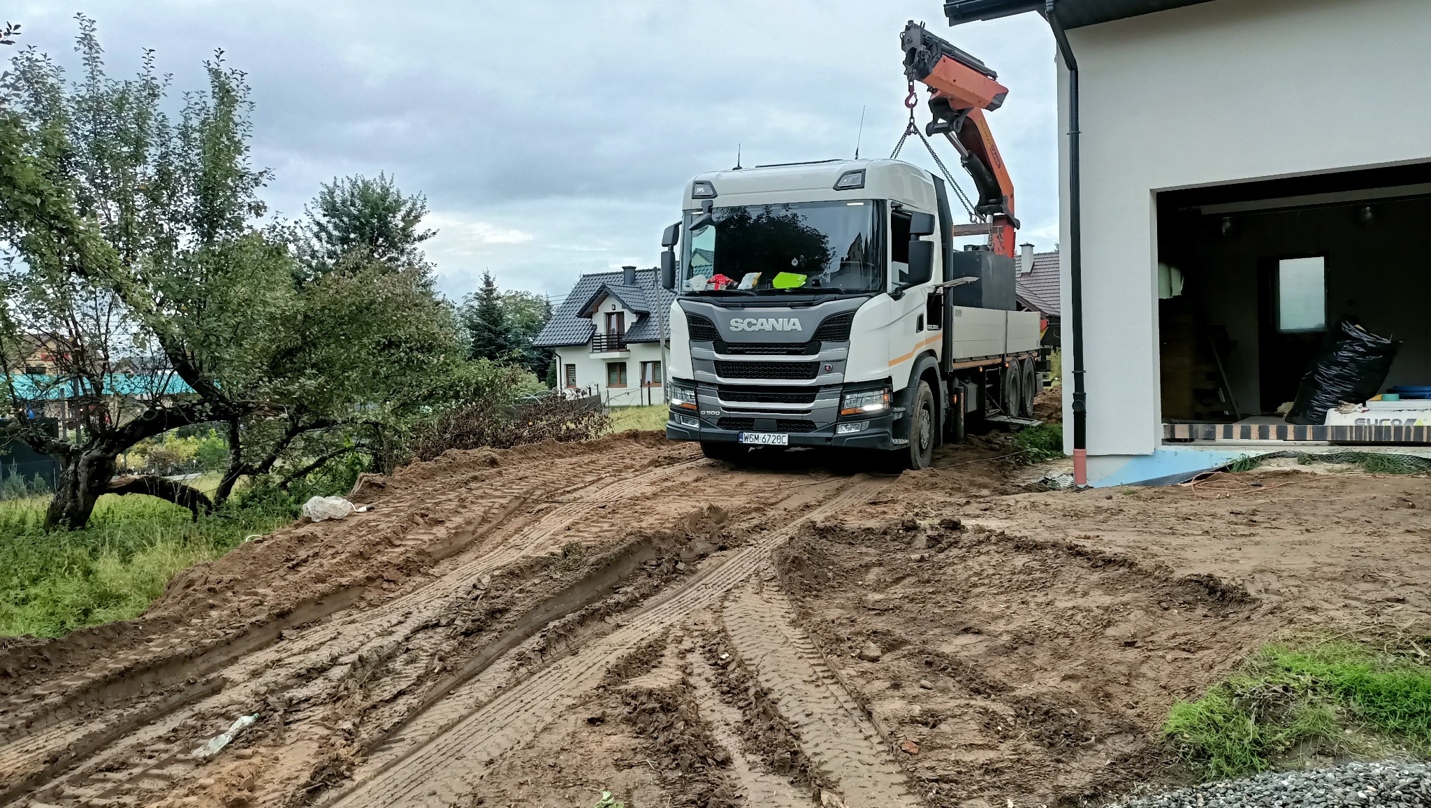Ciężarówka Scania z dźwigiem na błotnistym terenie budowy, obok biały budynek w trakcie prac. Ślady opon na ziemi. W tle widoczny dom jednorodzinny i drzewa.