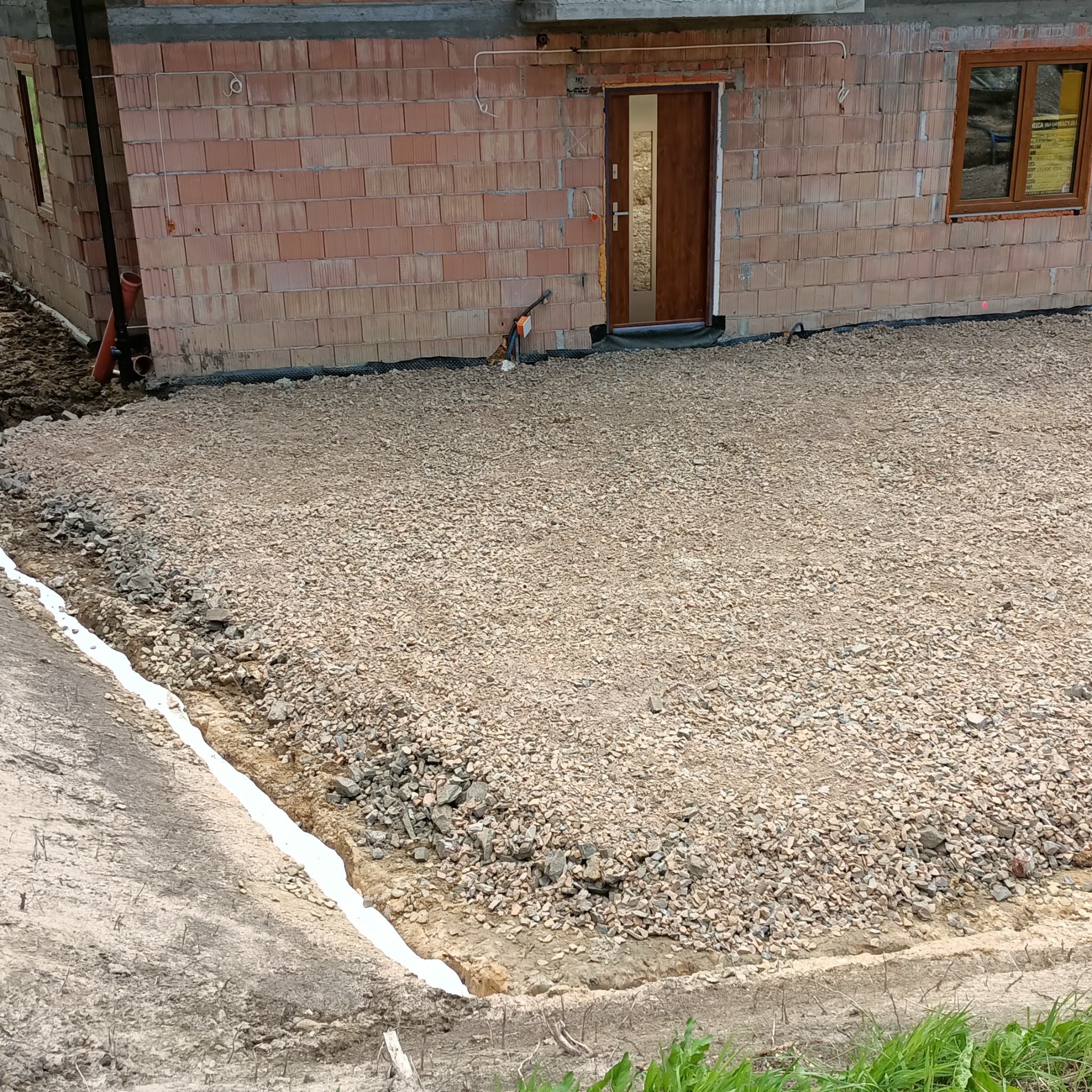 Przygotowany teren pod budowę domu z widoczną warstwą utwardzonego kruszywa i fundamentami w wykopie. Widoczny fragment budynku z cegły.