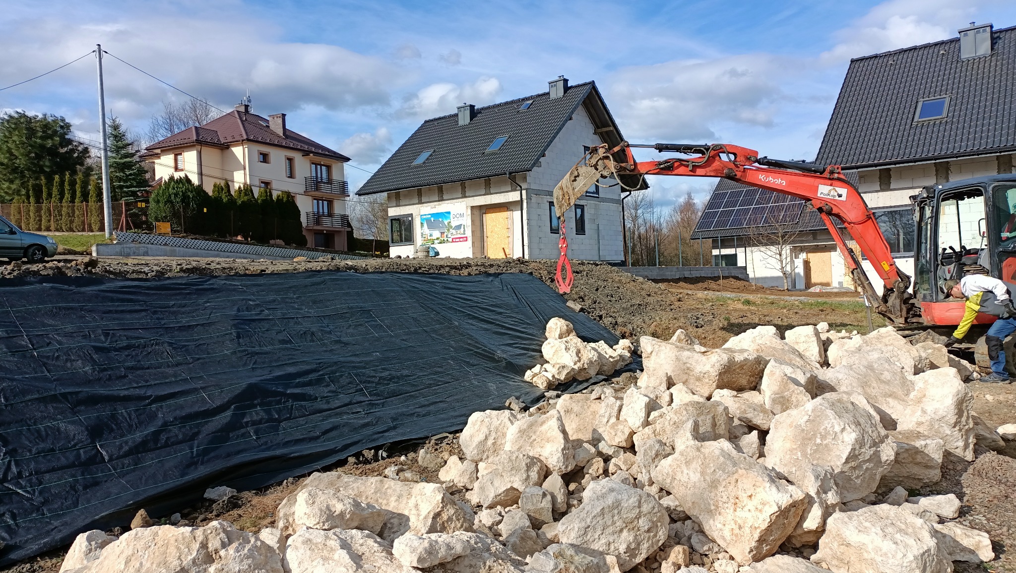 Czerwona koparka Kubota na placu budowy, obok sterta jasnych kamieni i czarna geowłóknina. W tle domy w różnym stanie ukończenia i błękitne niebo.