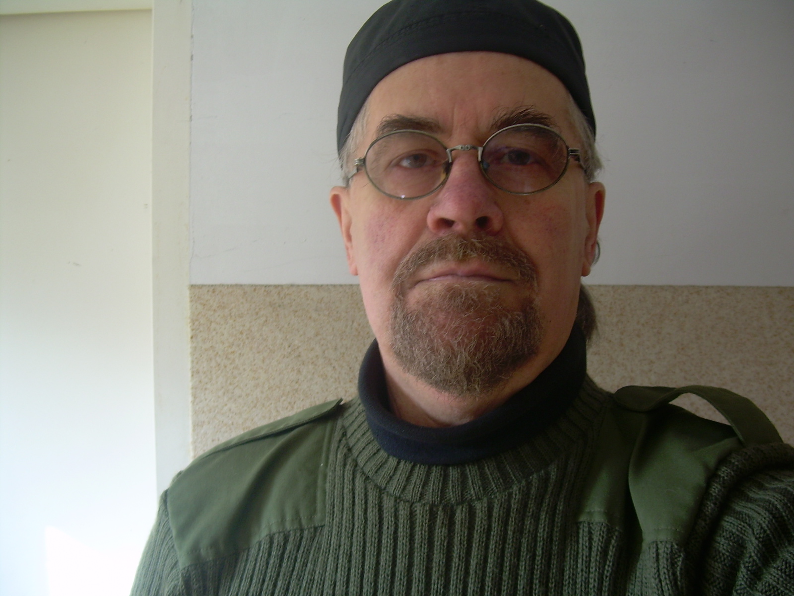 Portret mężczyzny z brodą i okularami w czapce i zielonym swetrze na tle ściany. Widoczny fragment ramienia z torbą. Ujęcie z bliska.