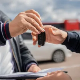 Przekazanie kluczyków do samochodu na tle białego auta i podpisanych dokumentów na czarnym podkładzie.