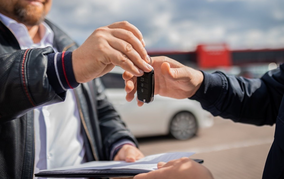 Przekazanie kluczyków do samochodu na tle białego auta i podpisanych dokumentów na czarnym podkładzie.