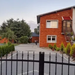 Dwupiętrowy dom z czerwonej cegły z szarymi ramami okiennymi, widoczny zza czarnego, kutego ogrodzenia, z motocyklem zaparkowanym przed garażem w tle.