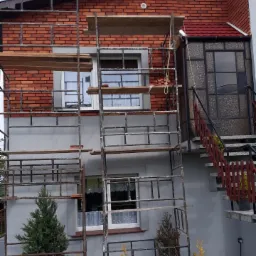 Dwukondygnacyjny dom w trakcie prac elewacyjnych, z widocznym rusztowaniem, świeżo wymurowaną elewacją z czerwonej cegły i fragmentem schodów wejściowych.