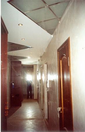 Korytarz w domu z nietypowym sufitem podwieszanym z kwadratowymi elementami i wbudowanym oświetleniem, widoczne drzwi z ciemnego drewna i ściany pokryte dekoracyjnym tynkiem.