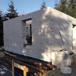 Wznoszenie ścian domu z bloczków betonu komórkowego, widoczne otwory okienne i drzwiowe, przygotowane drewno i narzędzia budowlane na placu budowy.