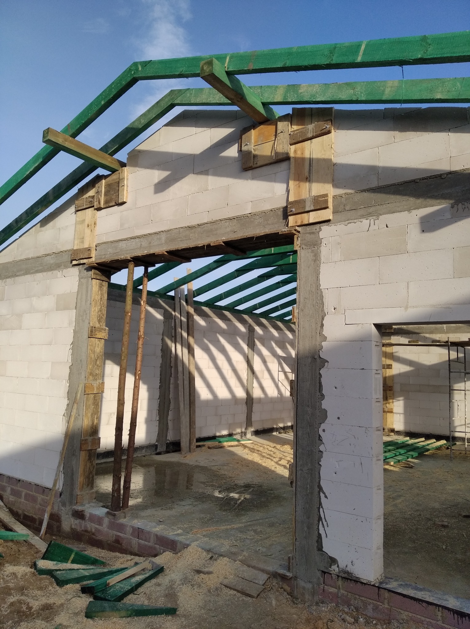 Surowy stan budowy domu z betonu komórkowego: ściany z otworami okiennymi i drzwiowymi, drewniana konstrukcja dachu pomalowana na zielono, drewniane stemple podtrzymujące nadproże, widoczne ślady...